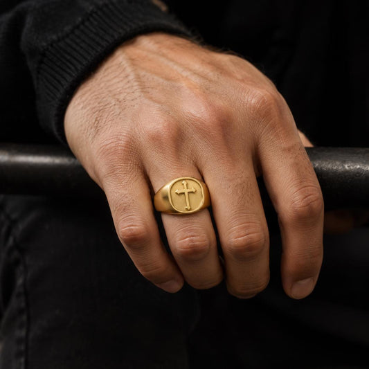 A close-up of a person's hand wearing a gold ring with a cross symbol on it.