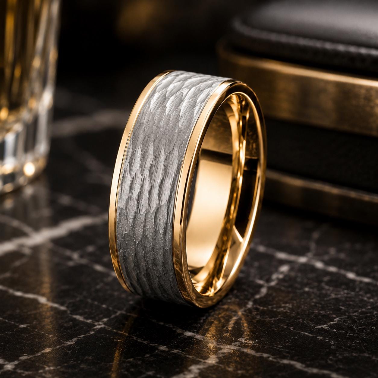 A gold and silver ring with a textured band sits on a black surface, with a glass of what appears to be a beverage in the background.