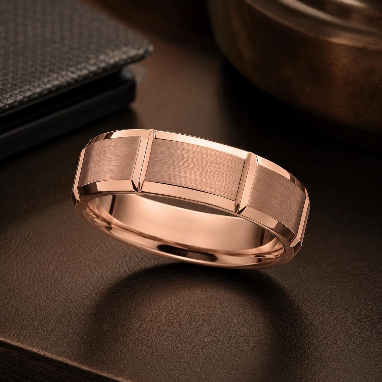A rose gold ring with a textured band is displayed on a dark surface, with a black wallet and a metal container visible in the background.