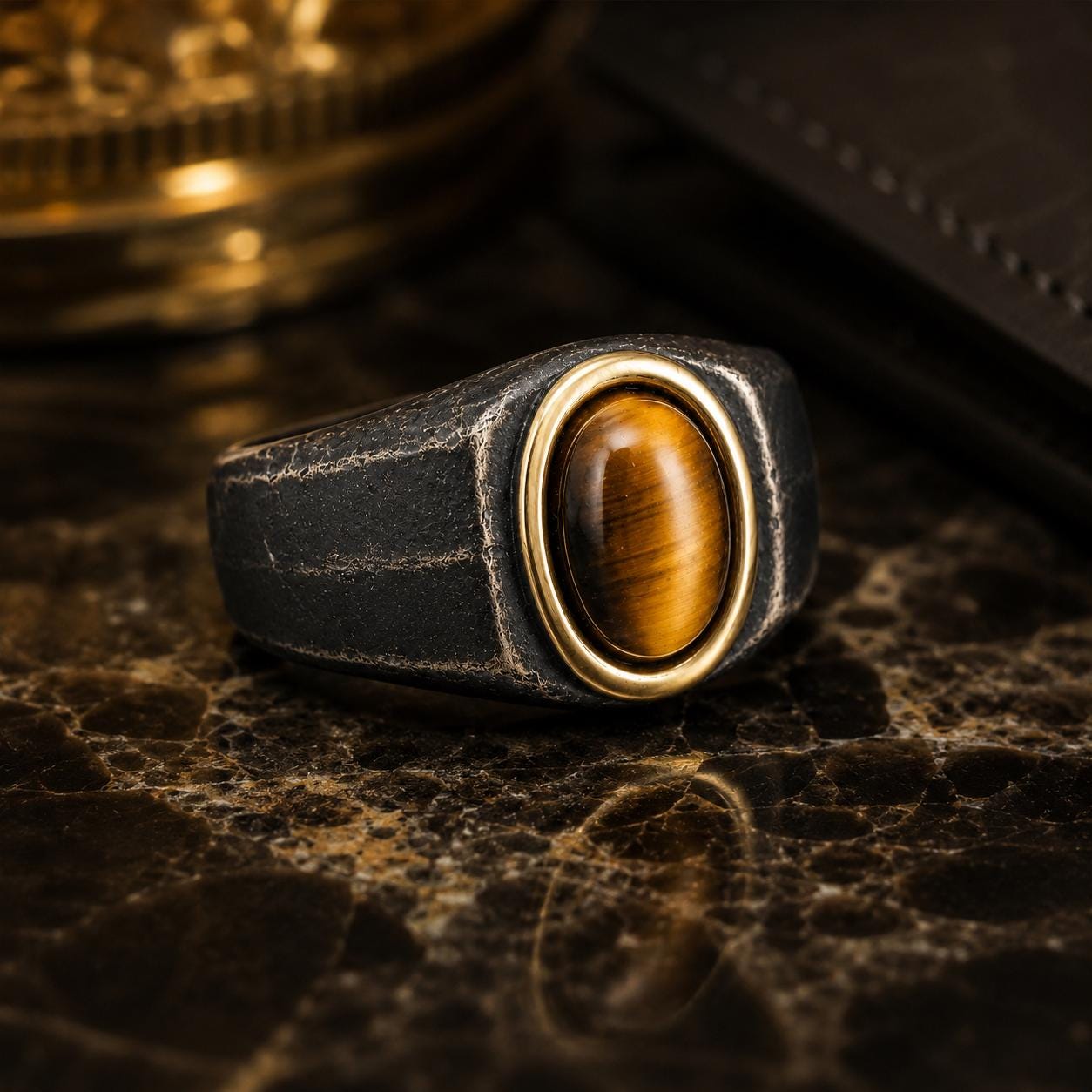 A close-up image of a ring with a large oval-shaped gemstone, possibly a tiger eye, set in a black metal band with a gold accent.