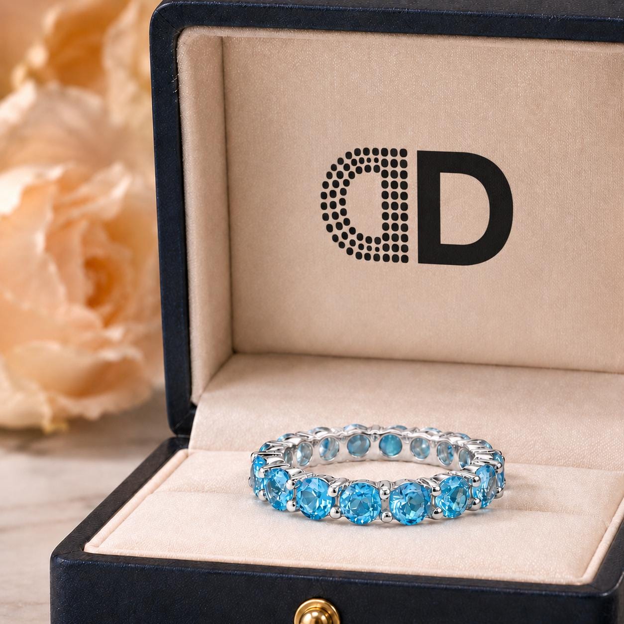 A blue gemstone ring is displayed in a black jewelry box with a logo on the lid.