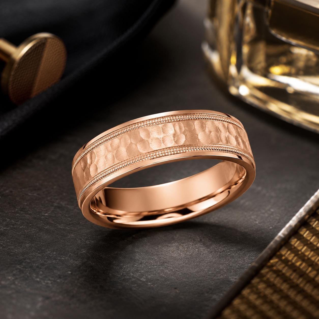 A gold-colored ring with a textured band is displayed on a dark surface, with a glass of what appears to be a golden liquid in the background.