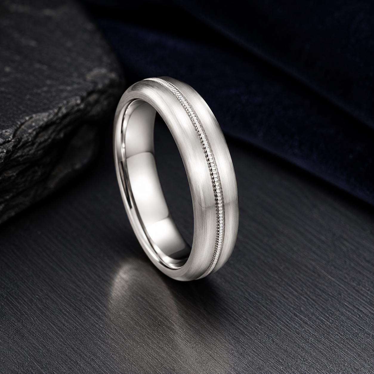 A silver ring with a unique design sits on a dark surface, surrounded by a dark background.