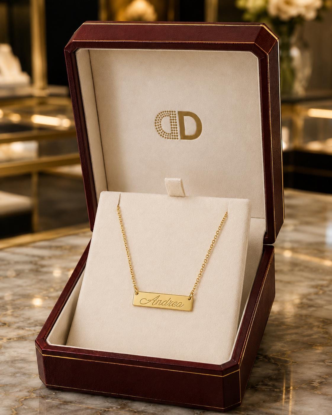 A gold necklace with a personalized nameplate is displayed in a burgundy jewelry box on a marble countertop.