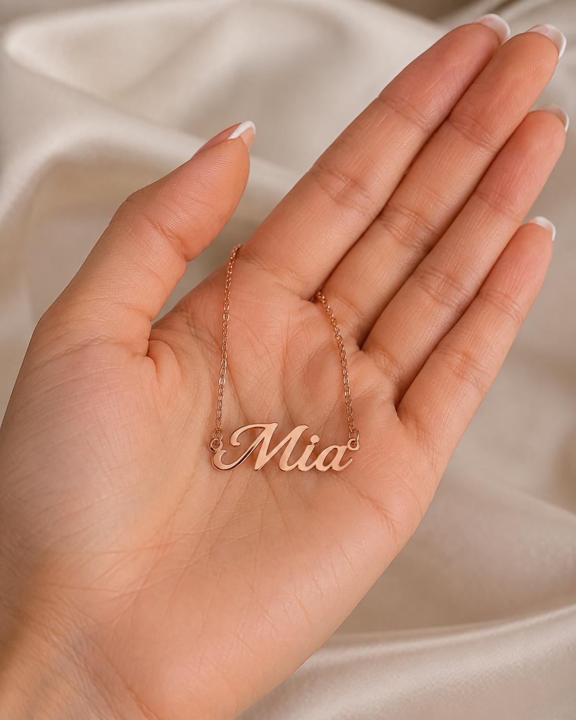 A hand holding a necklace with the name "Mia" engraved on it.