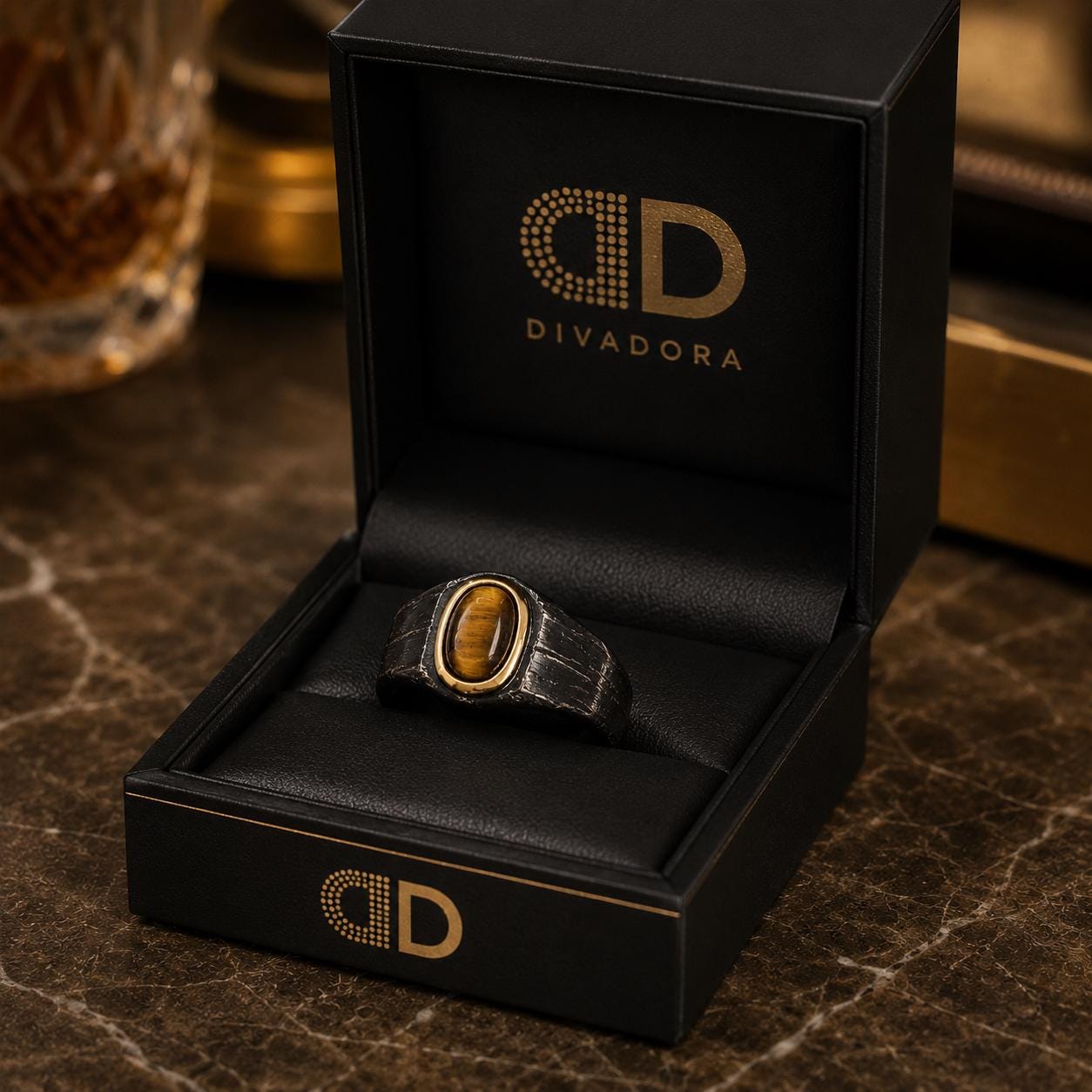 A black jewelry box with a gold logo and a ring inside it, placed on a marble surface.