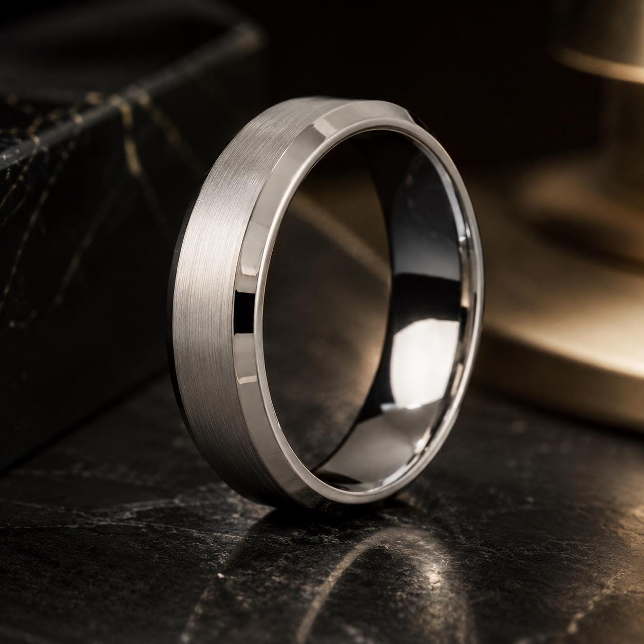 A silver ring with a smooth, polished surface sits on a dark surface, reflecting light and creating a shiny appearance.