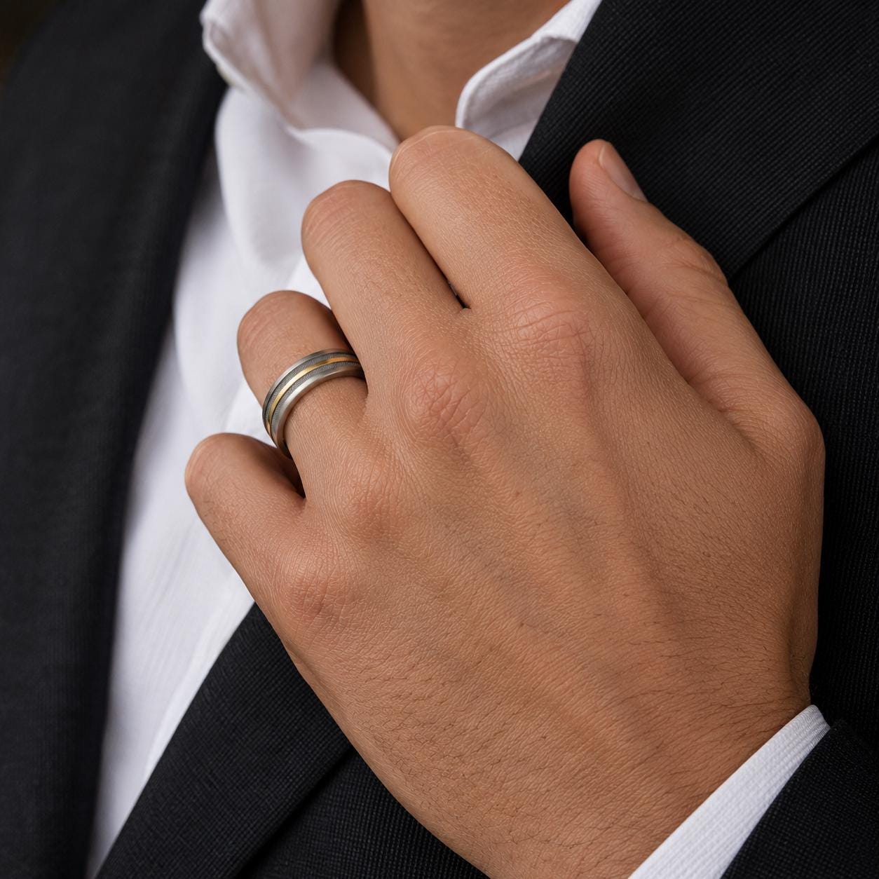 A close-up image of a person's hand wearing a wedding ring, with a black suit jacket visible.