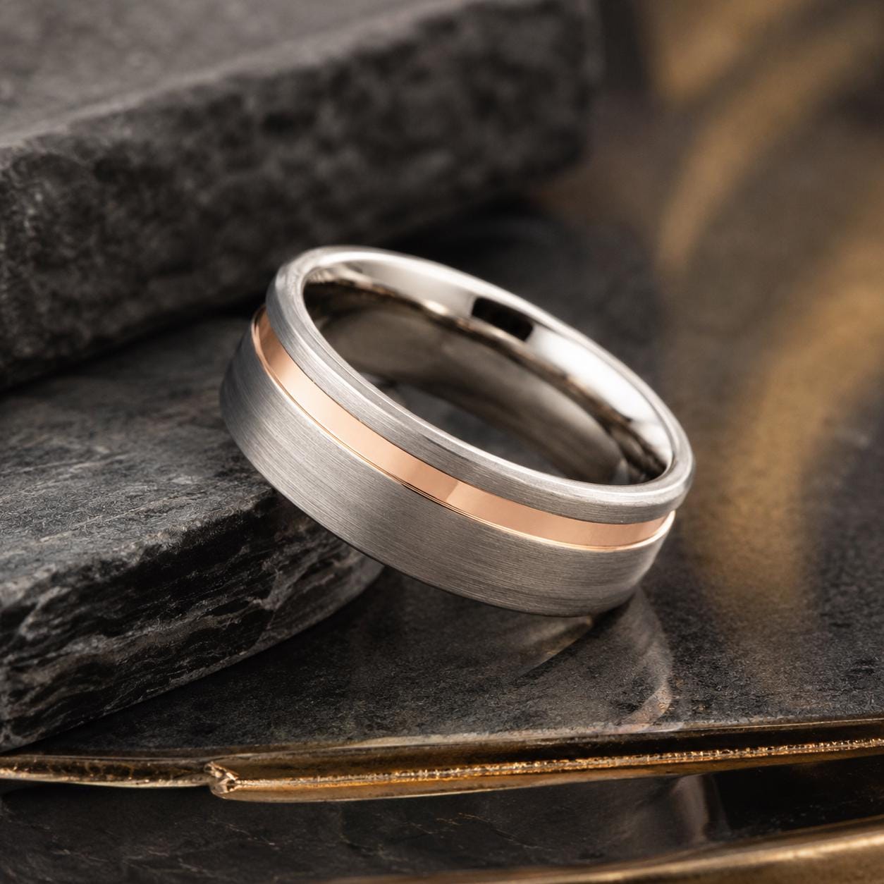 A silver and gold wedding band with a thin gold stripe is displayed on a black surface, with a blurred background.