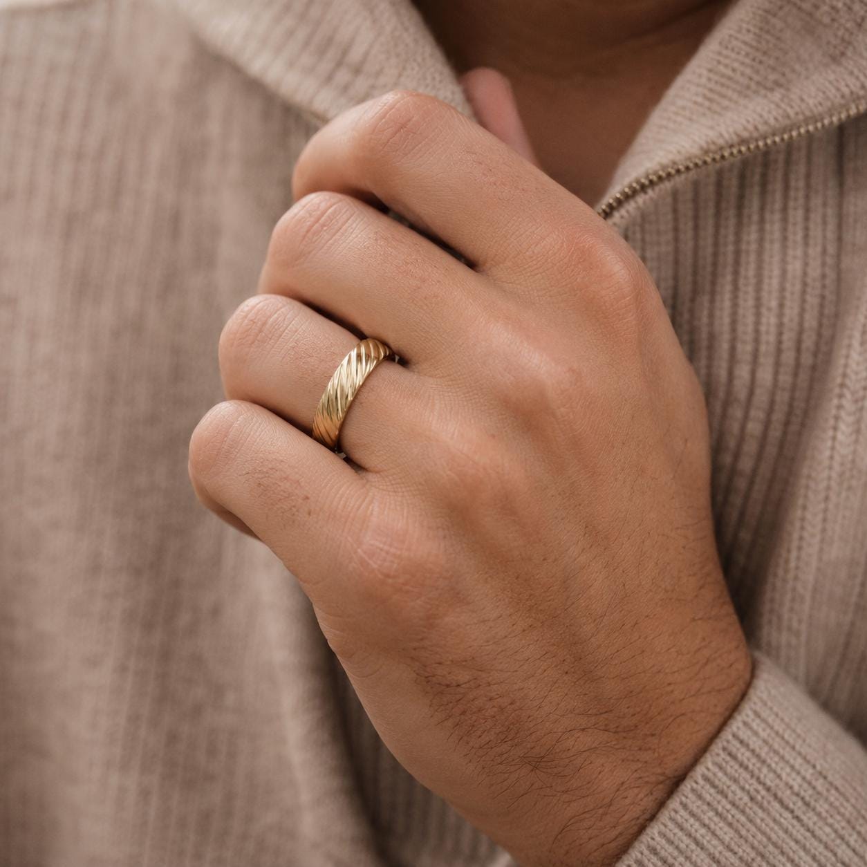 A close-up of a person's hand wearing a gold ring, with a beige sweater partially visible in the background.