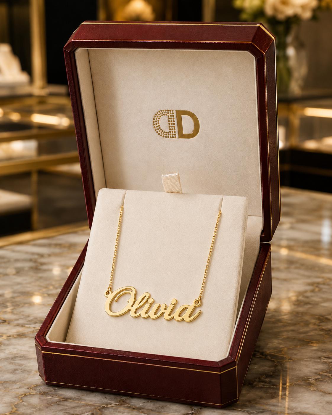 A gold necklace with the name "Olivia" engraved on it, displayed in a burgundy jewelry box with a gold logo.