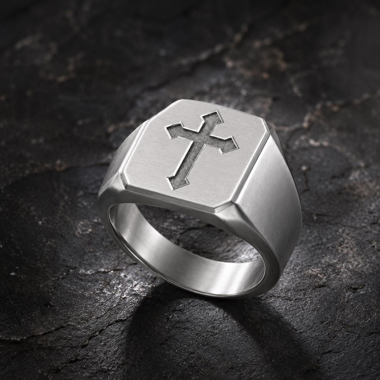 A silver ring with a cross design on it, resting on a dark, textured surface.