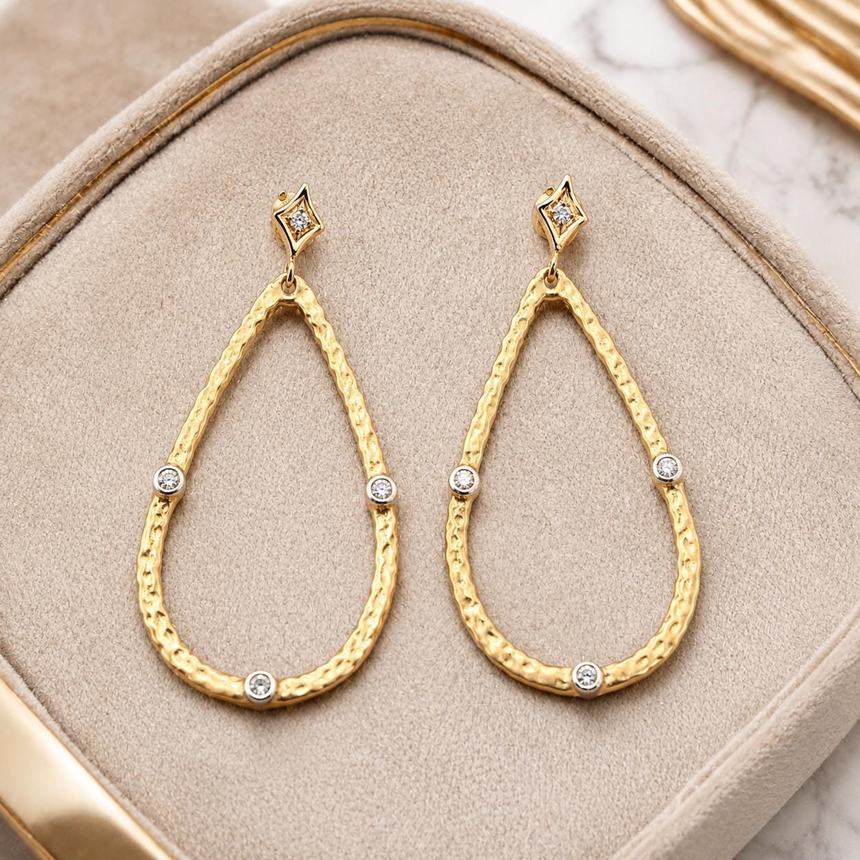 A pair of gold earrings with diamond accents, displayed on a beige velvet background.