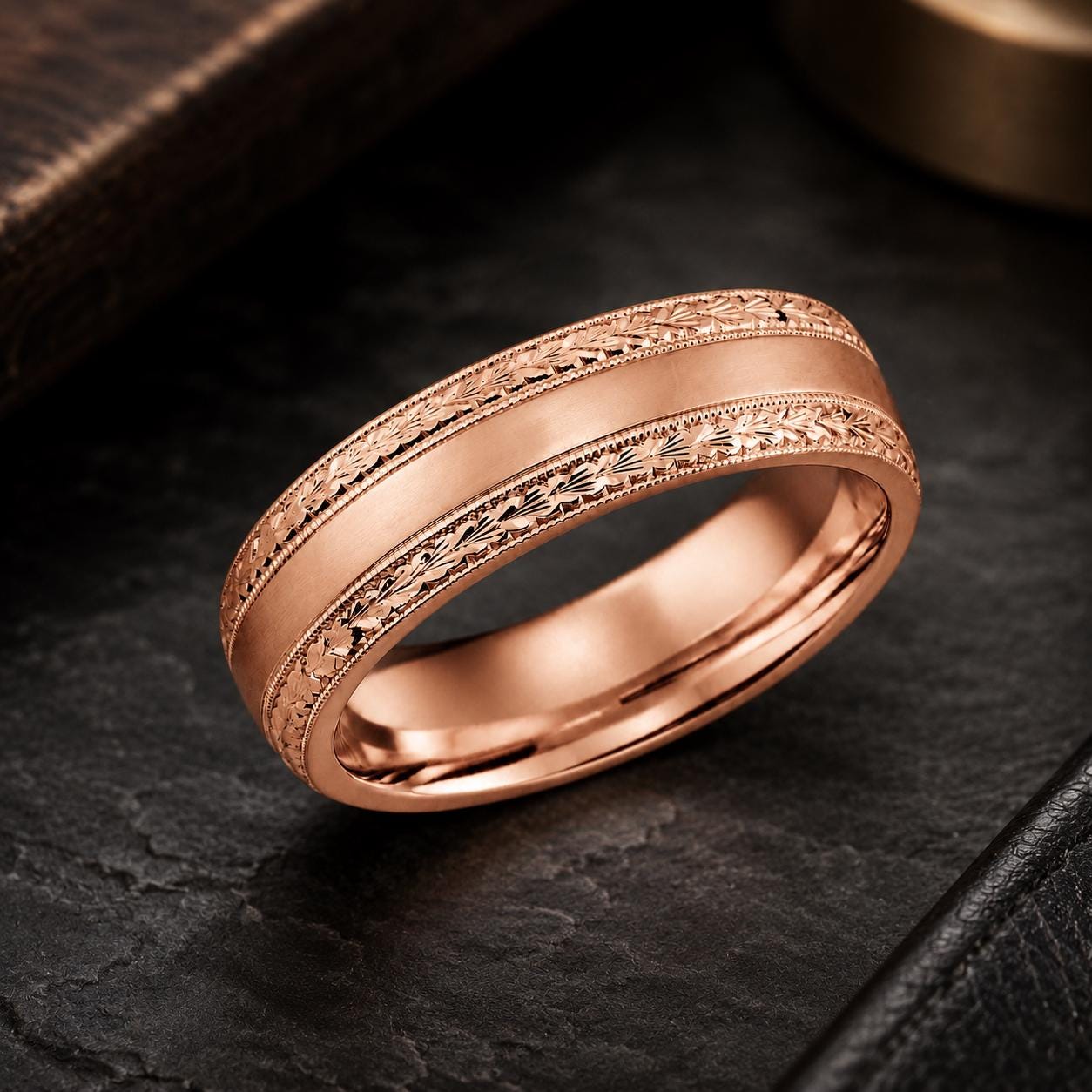 A rose gold wedding band with an engraved pattern on the band and a dark background.