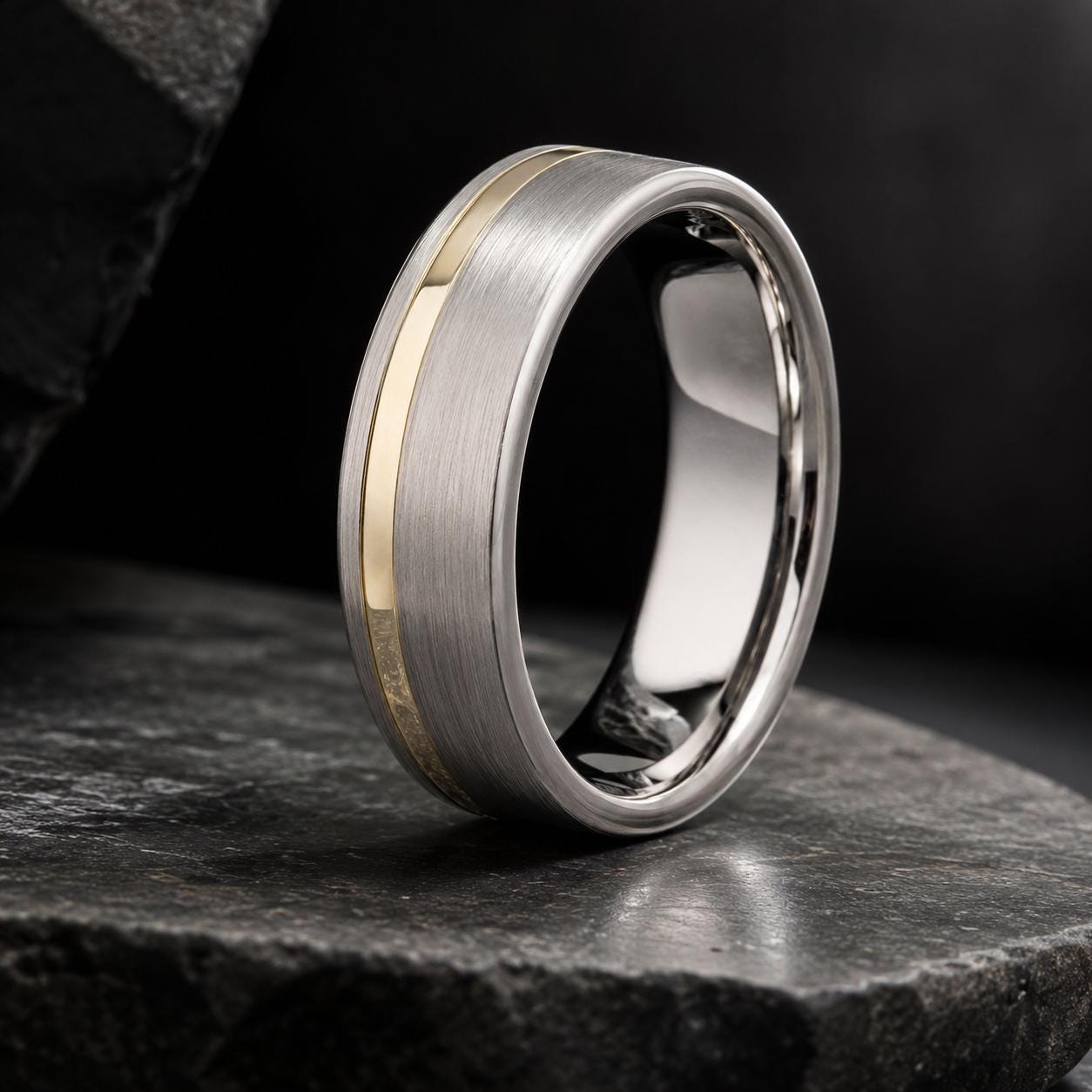 A silver and gold wedding band with a thin band and a thicker band with a reflective surface, resting on a dark surface.