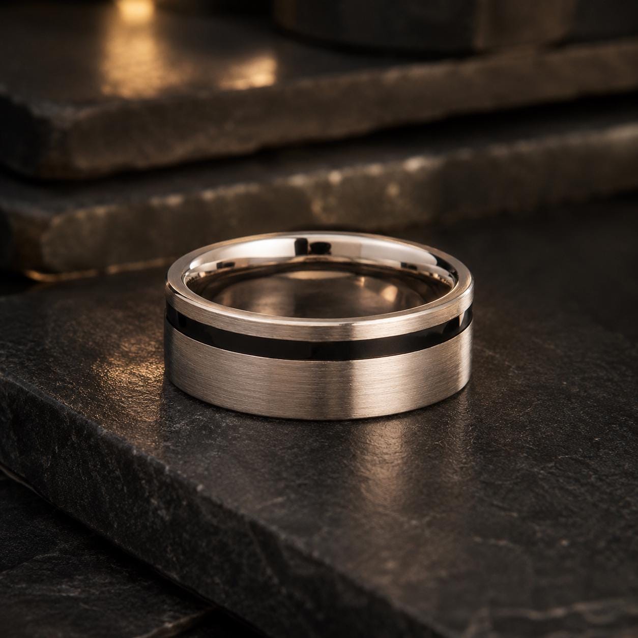 A silver ring with a black stripe is placed on a dark surface, possibly a stone or a black table.