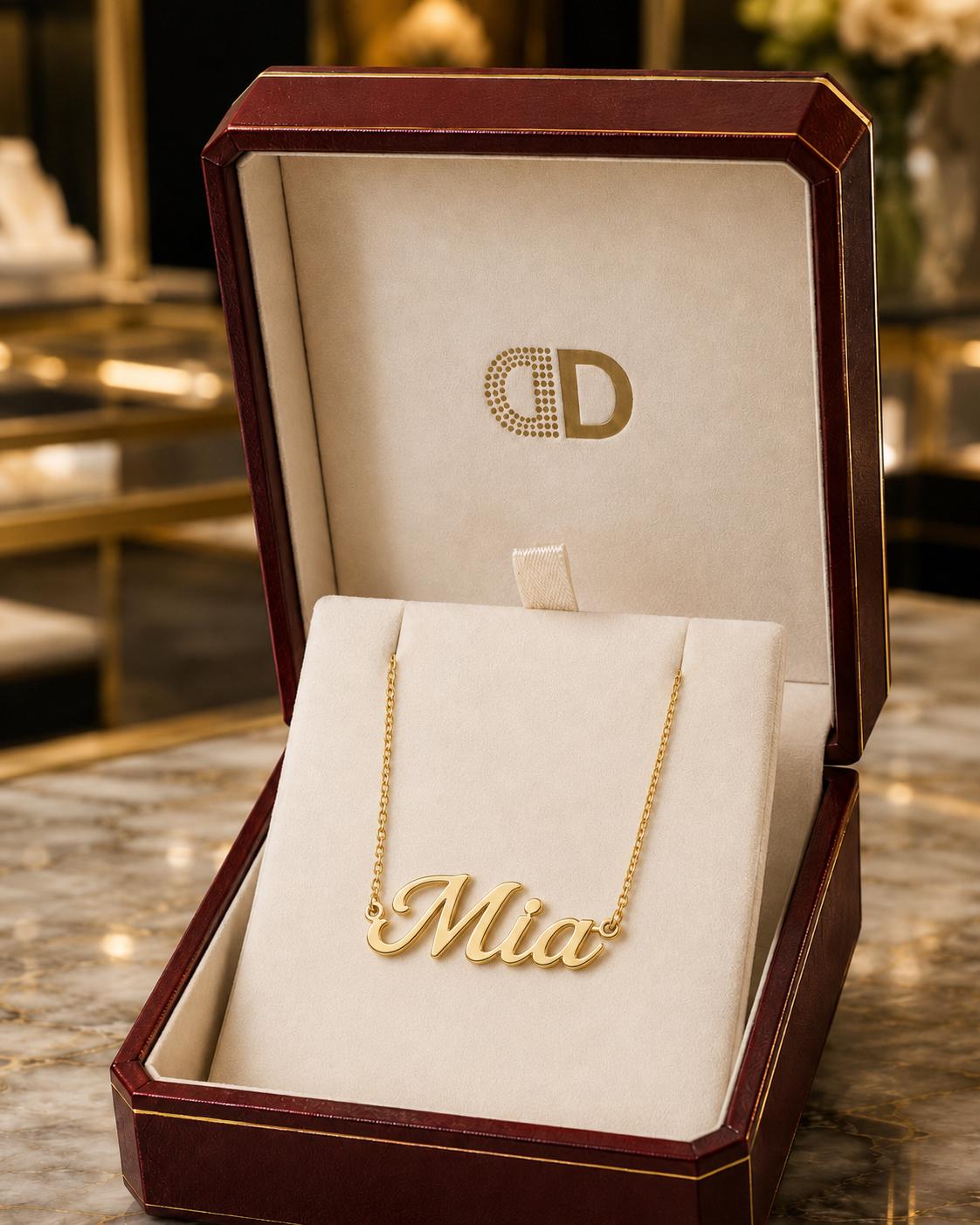 A gold necklace with the name "Mia" engraved on it, displayed in a red jewelry box with a gold logo.