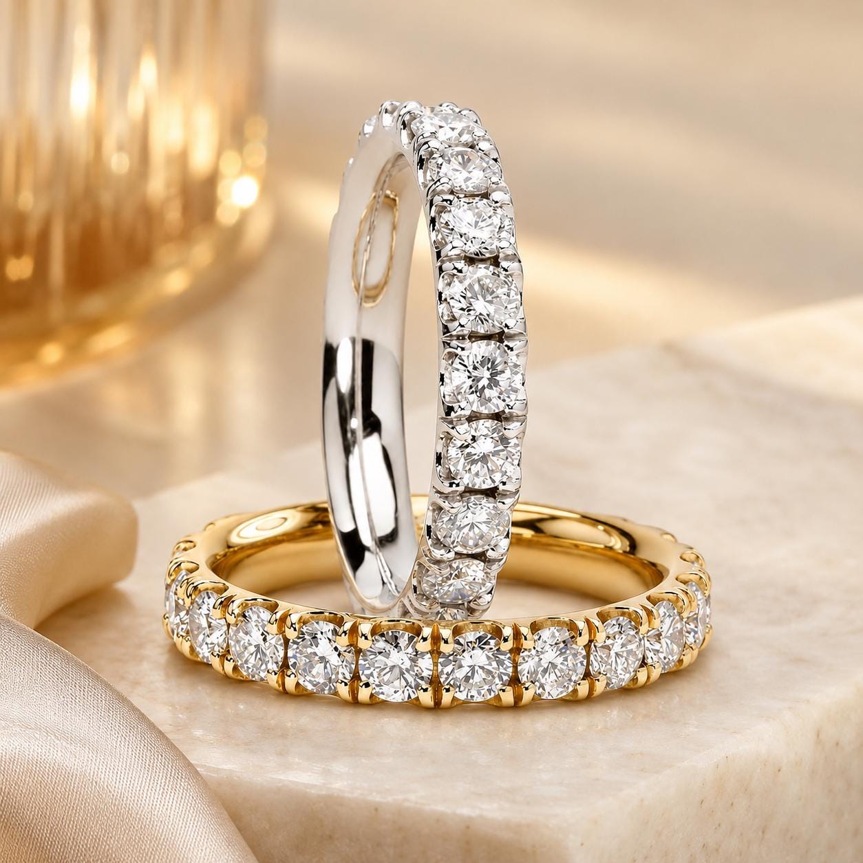 Two diamond-encrusted rings are displayed on a marble surface, one ring is a gold band with a row of diamonds, and the other is a silver band with a row of diamonds.