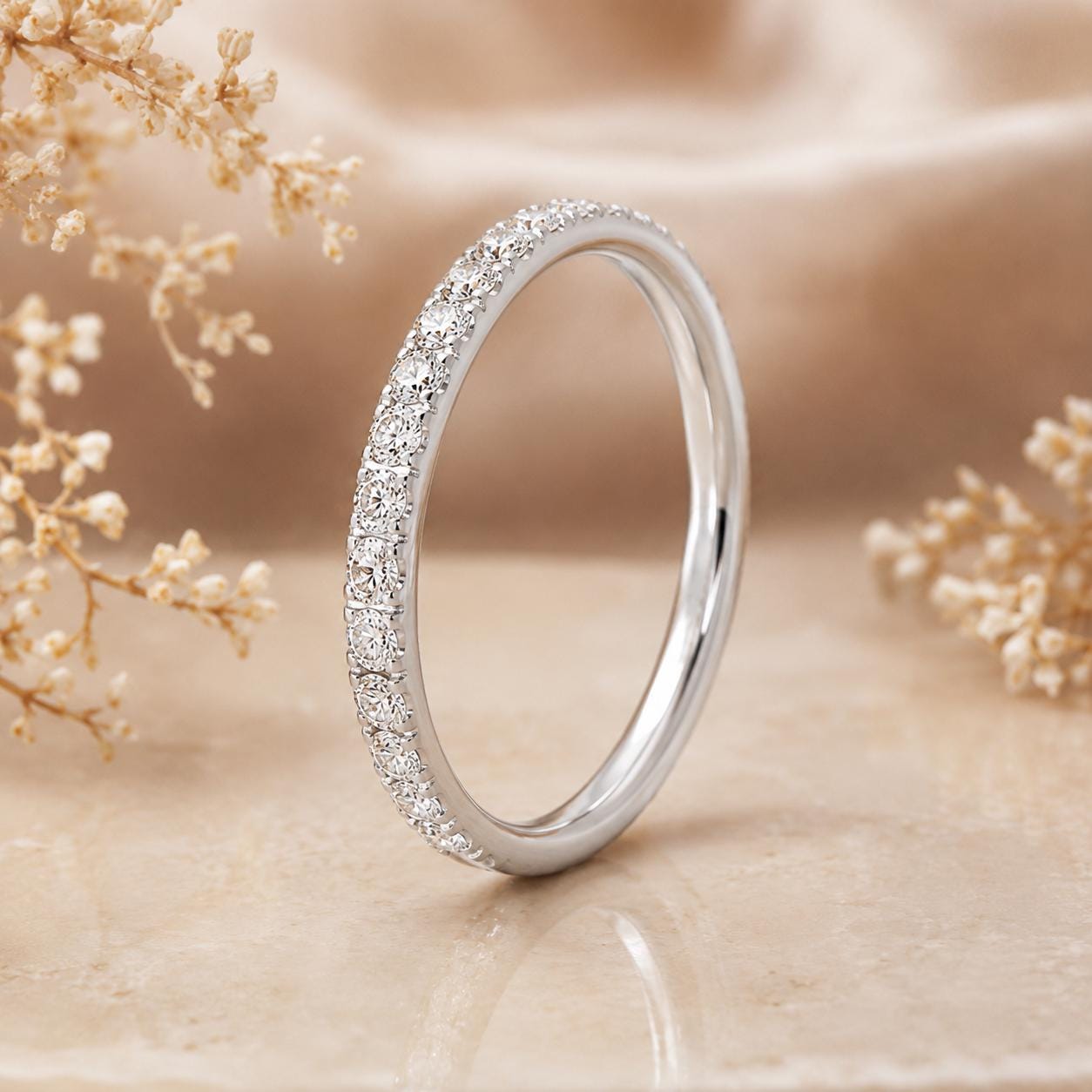 A silver band ring with a row of small diamonds set in the center, placed on a beige surface with delicate white flowers in the background.