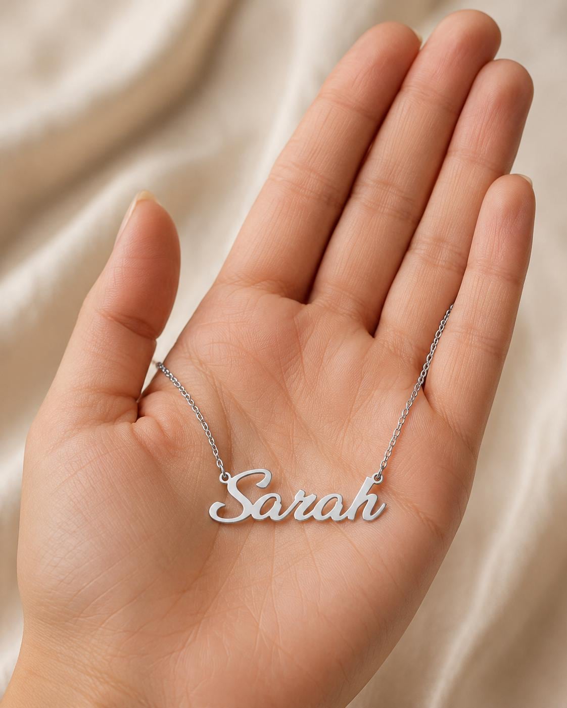 A hand holding a silver necklace with the name "Sara" engraved on it.