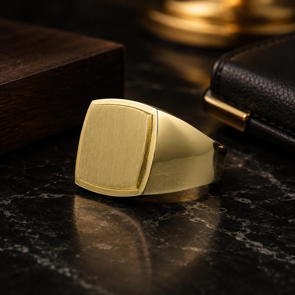 a gold ring with a square cutout on a black marble surface, accompanied by a wooden box and a black leather wallet.