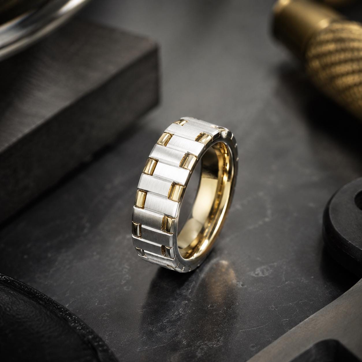 A silver and gold ring with a unique design sits on a black surface, surrounded by various tools and objects.