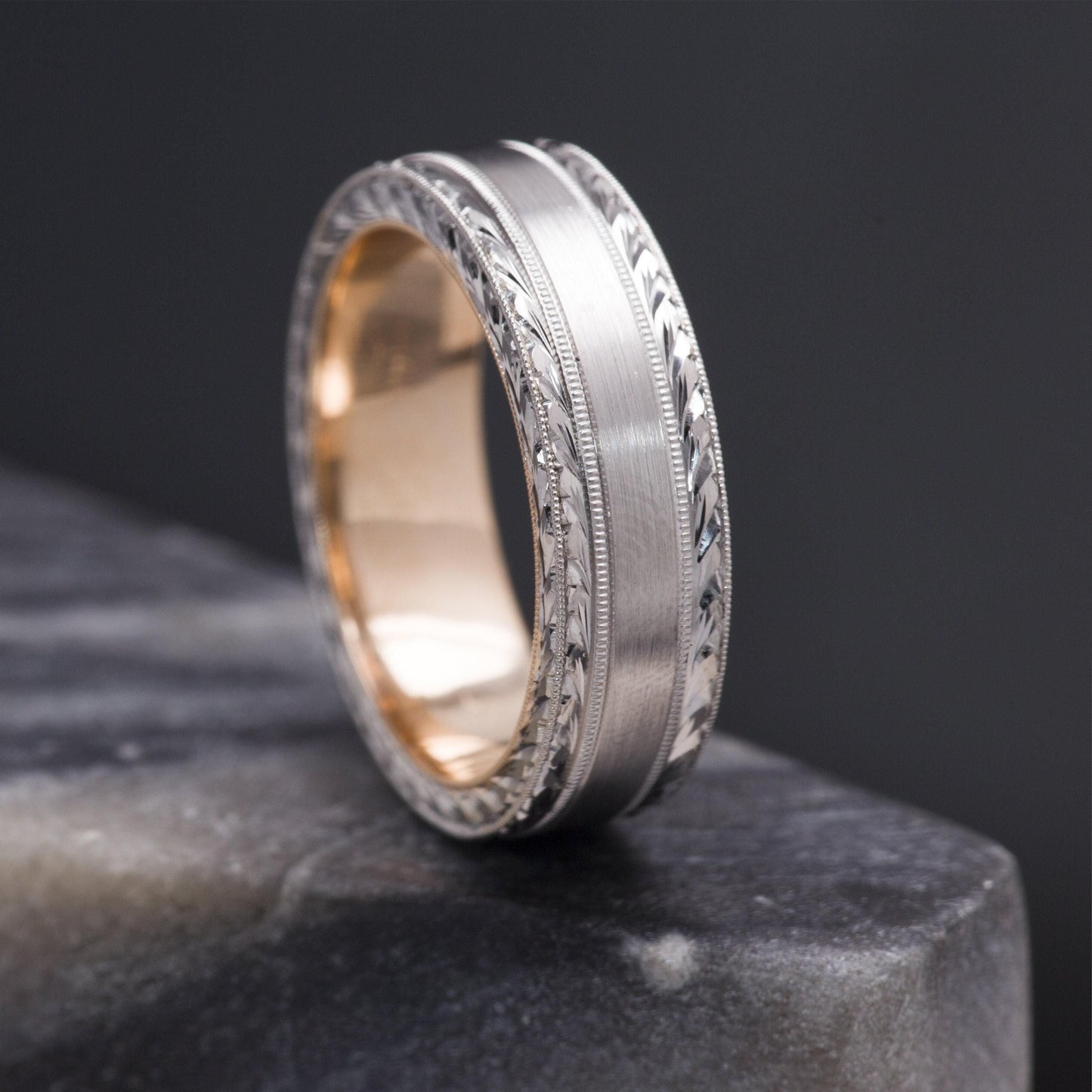 A silver ring with a gold band is displayed on a gray surface against a dark background.