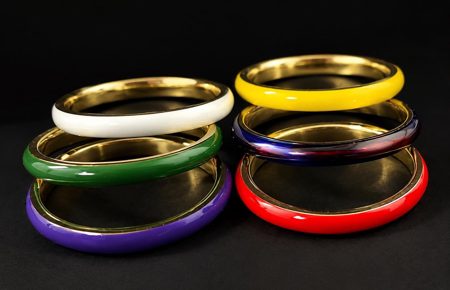 A collection of six colorful bracelets, including white, green, purple, red, blue, and yellow, arranged in a stack on a black background.