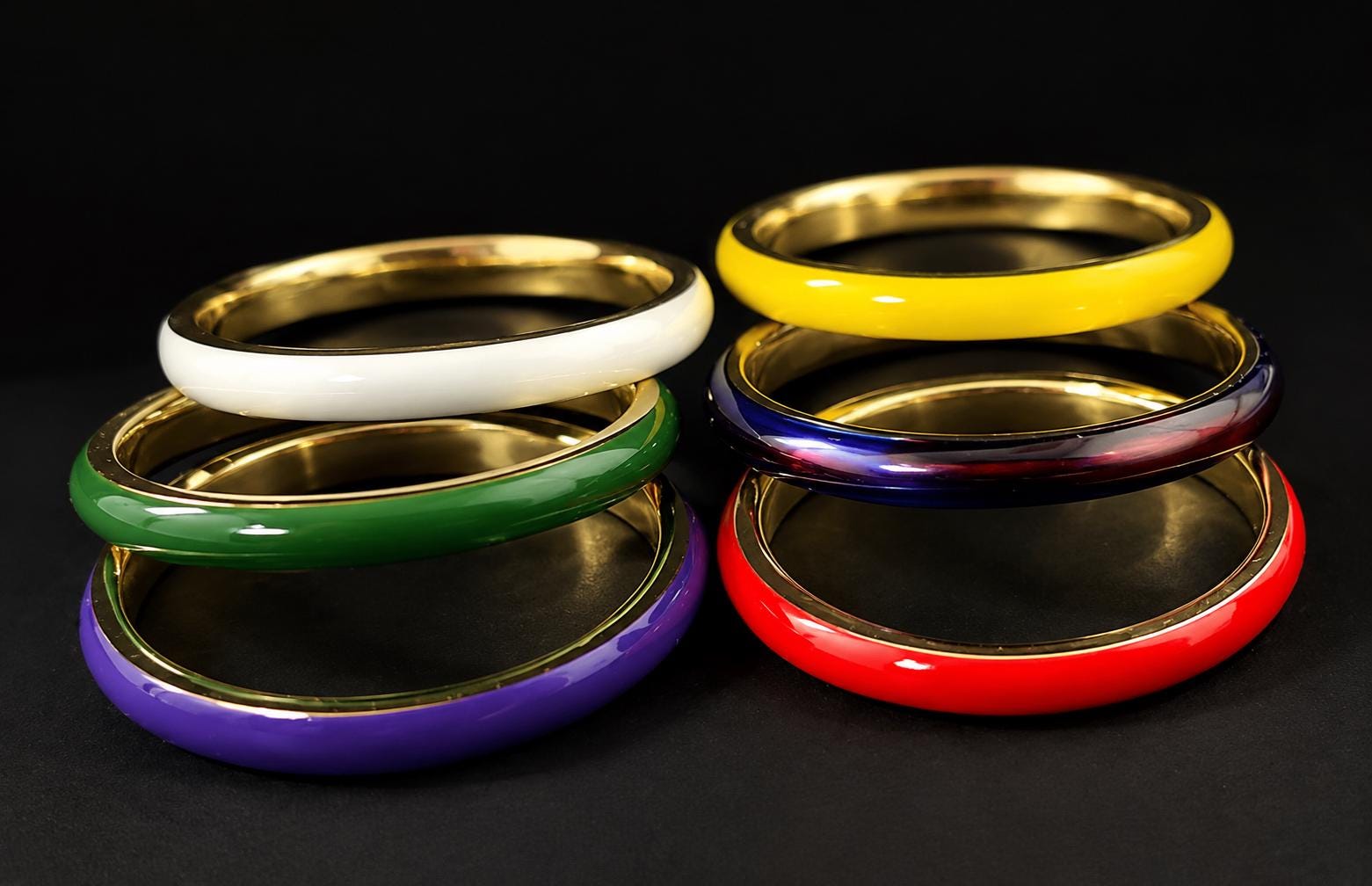 A collection of six colorful bracelets, including white, green, purple, red, blue, and yellow, arranged in a stack on a black background.