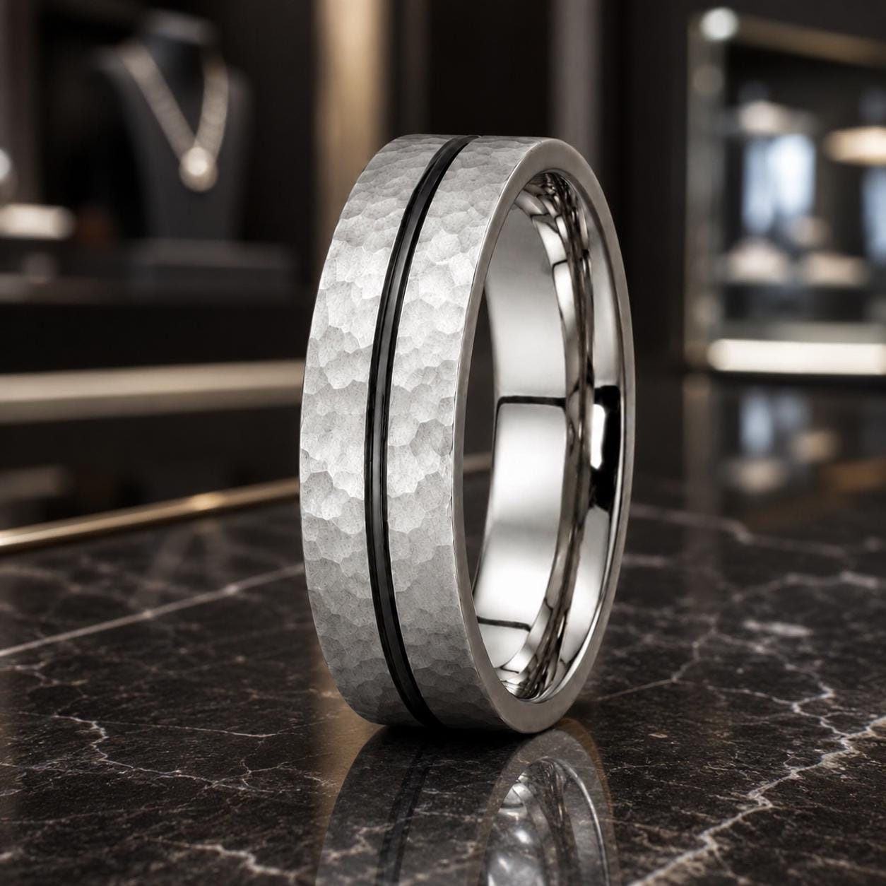 A silver ring with a textured surface and a thin black band is displayed on a marble countertop.