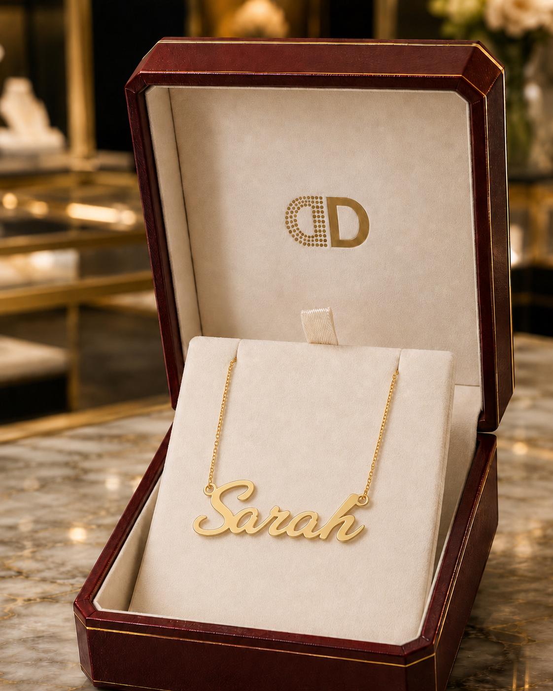 A gold necklace with the name "Sara" engraved on it, displayed in a red jewelry box with a gold-colored interior.