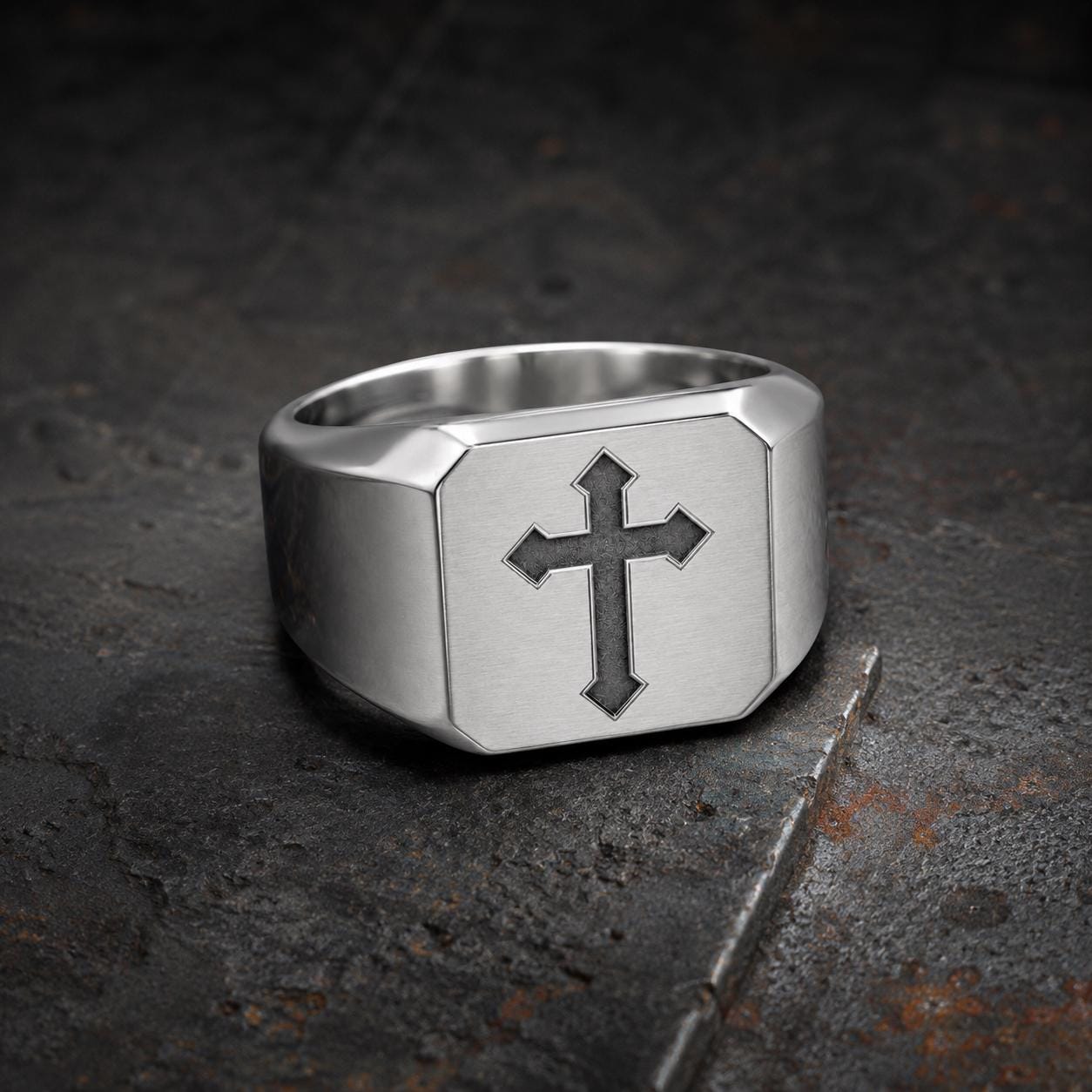 A silver ring with a cross symbol on it, placed on a dark, textured surface.