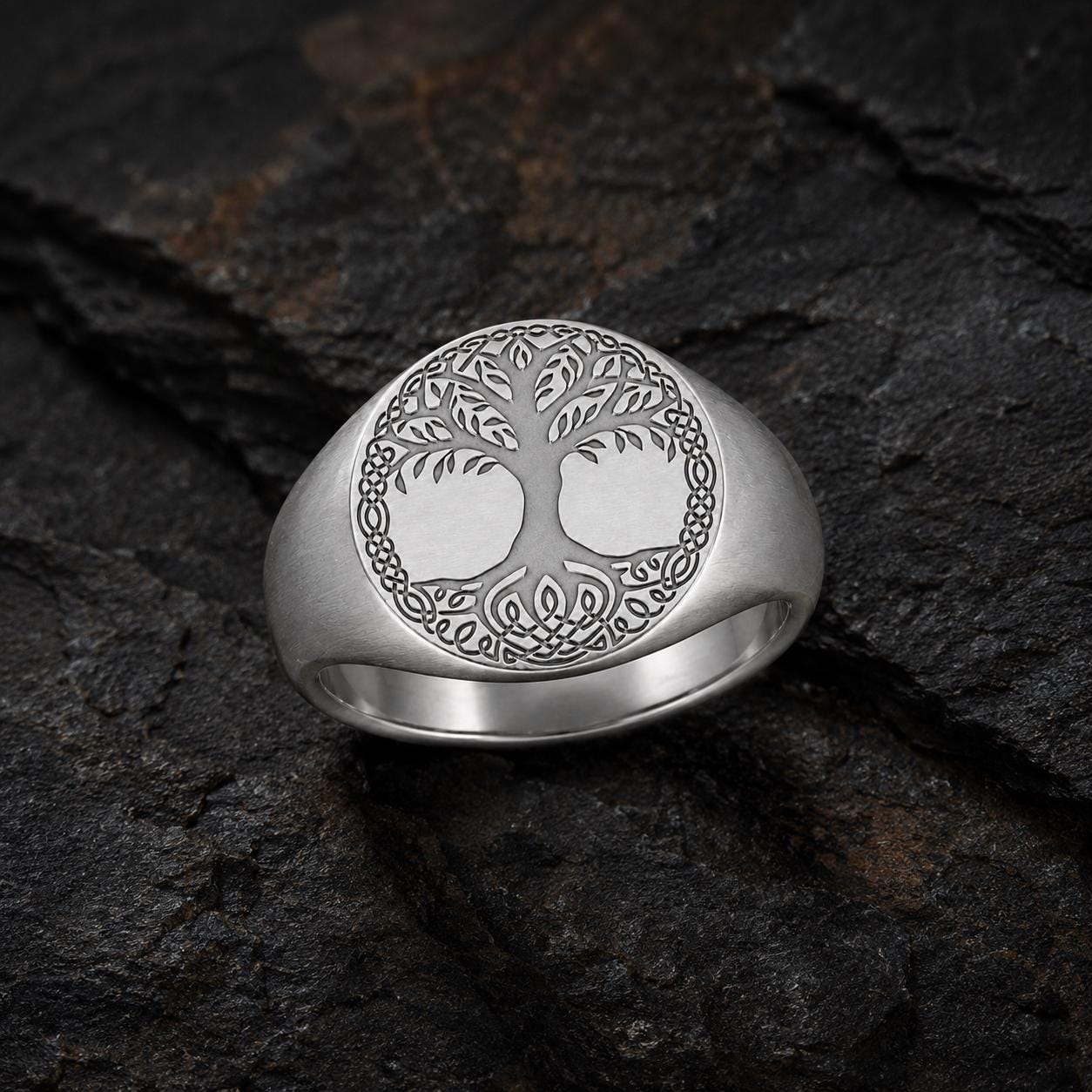 A silver ring with a tree design on it, resting on a dark surface.
