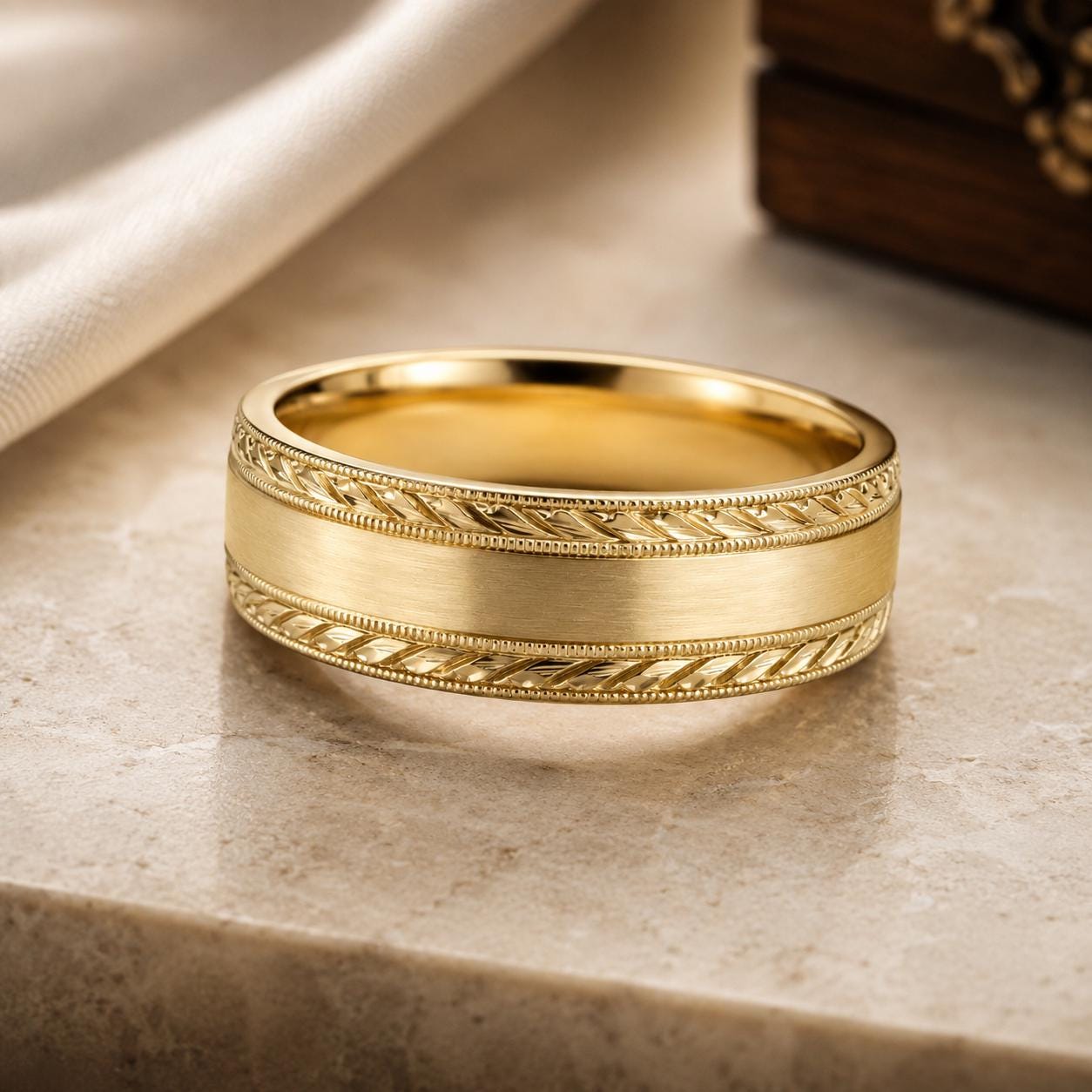 A gold wedding band with a decorative band design sits on a marble surface.