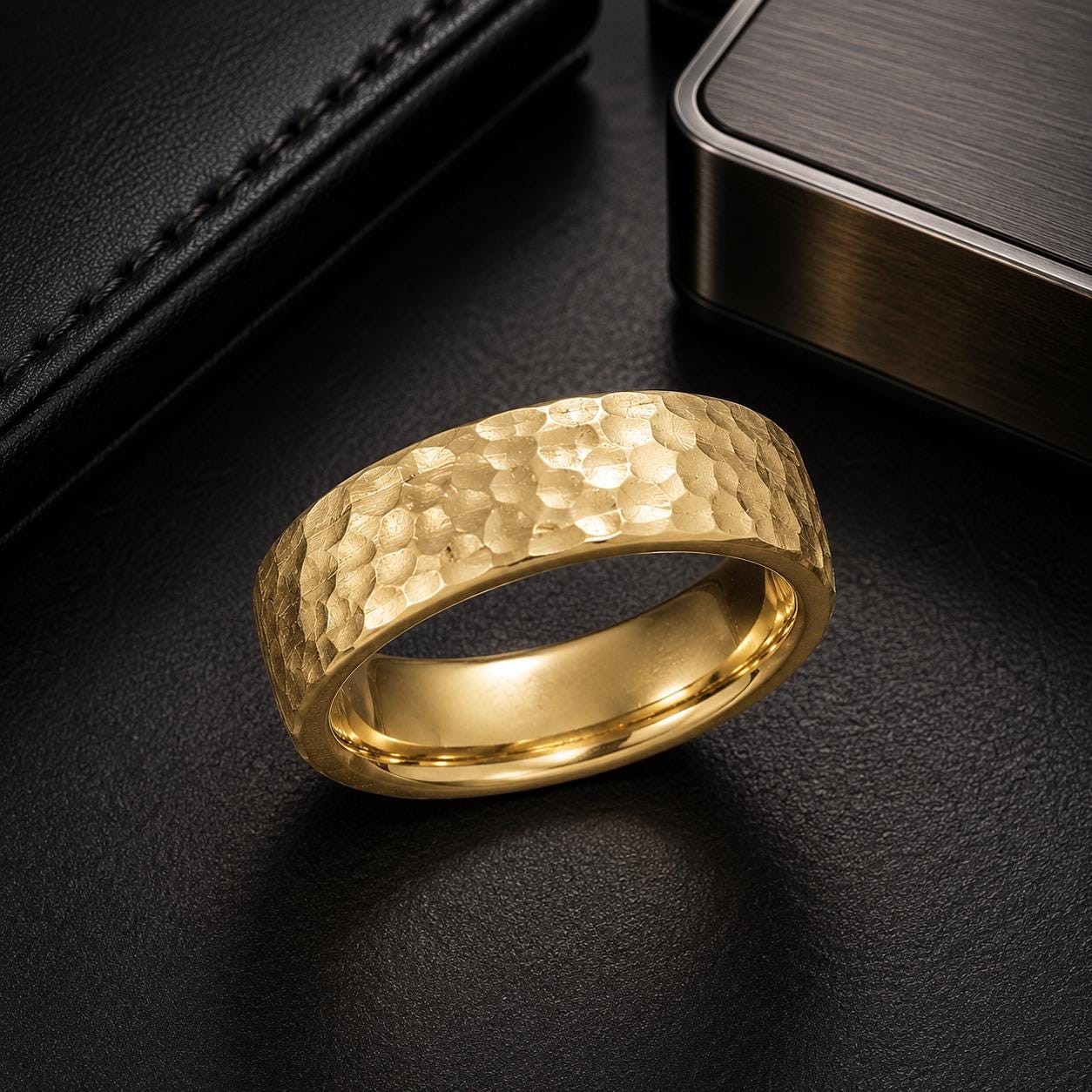 A gold ring with a textured surface sits on a black surface, surrounded by other objects such as a black leather wallet and a black box.