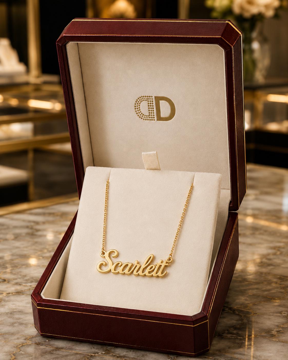A gold necklace with the name "Scarlett" engraved on it, displayed in a burgundy jewelry box with a gold logo.