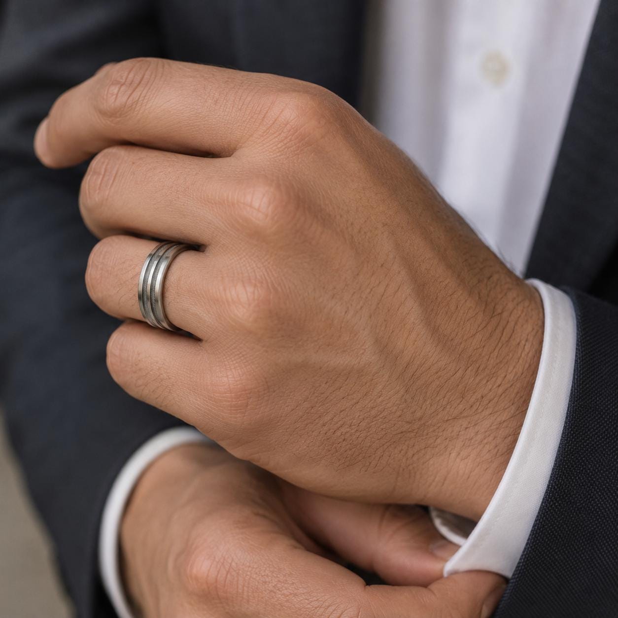 A close-up image of a person's hand wearing a ring, with a suit jacket visible in the background.