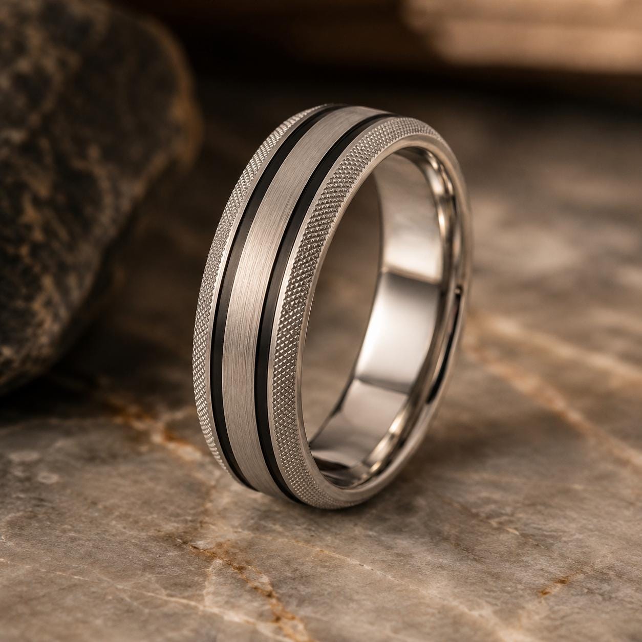 A silver ring with a textured band and a black stripe is displayed on a marble surface.