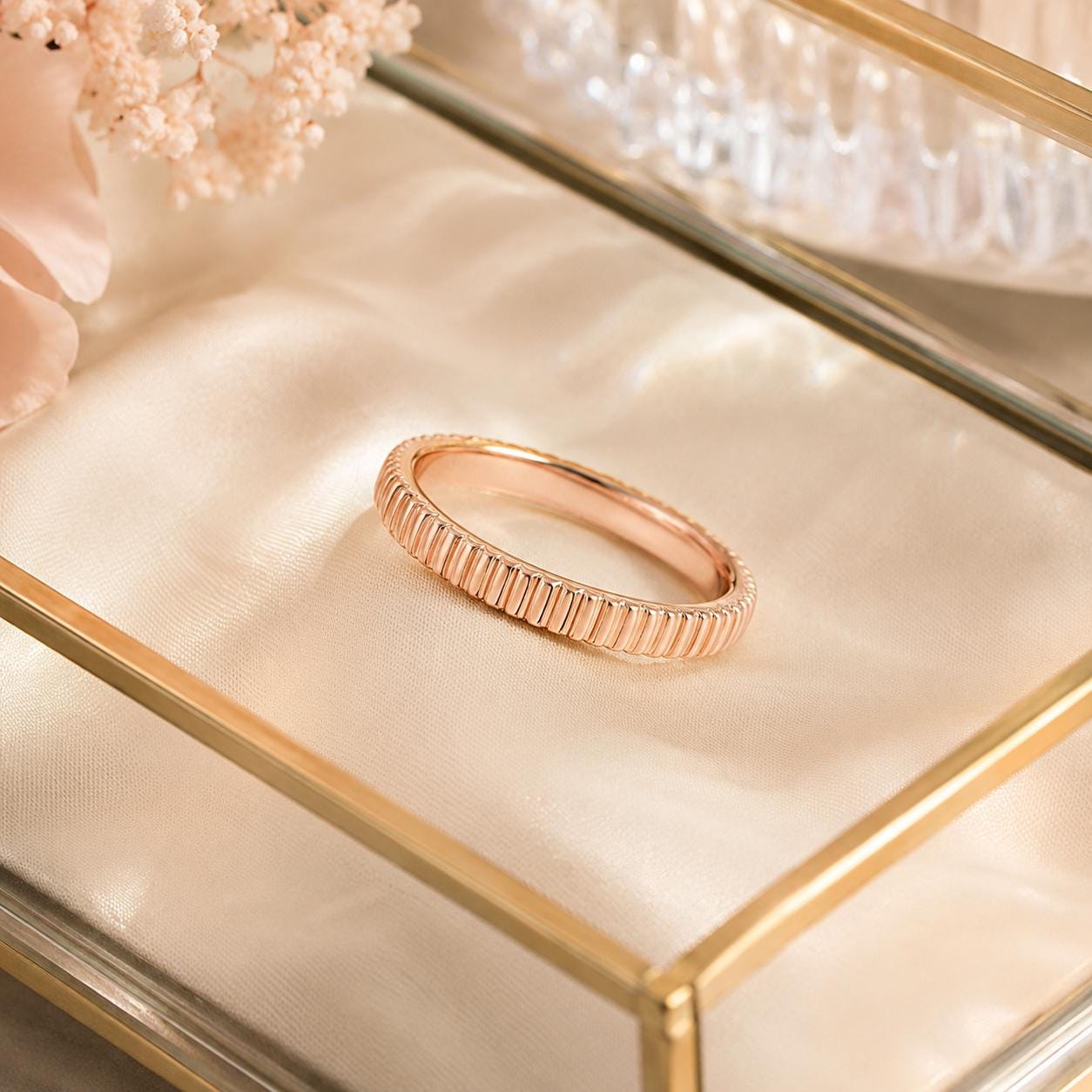 A gold ring with a textured band is displayed on a white fabric surface, with a glass container and pink flowers in the background.