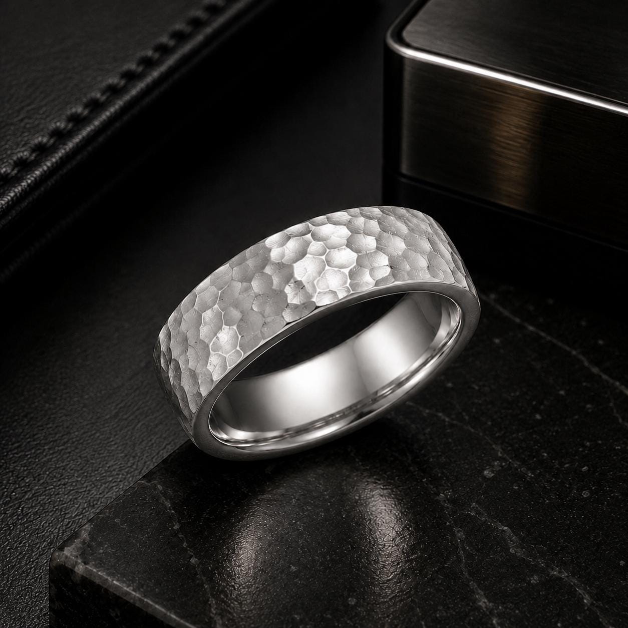 A silver ring with a textured surface sits on a black surface, surrounded by a black leather-like background.