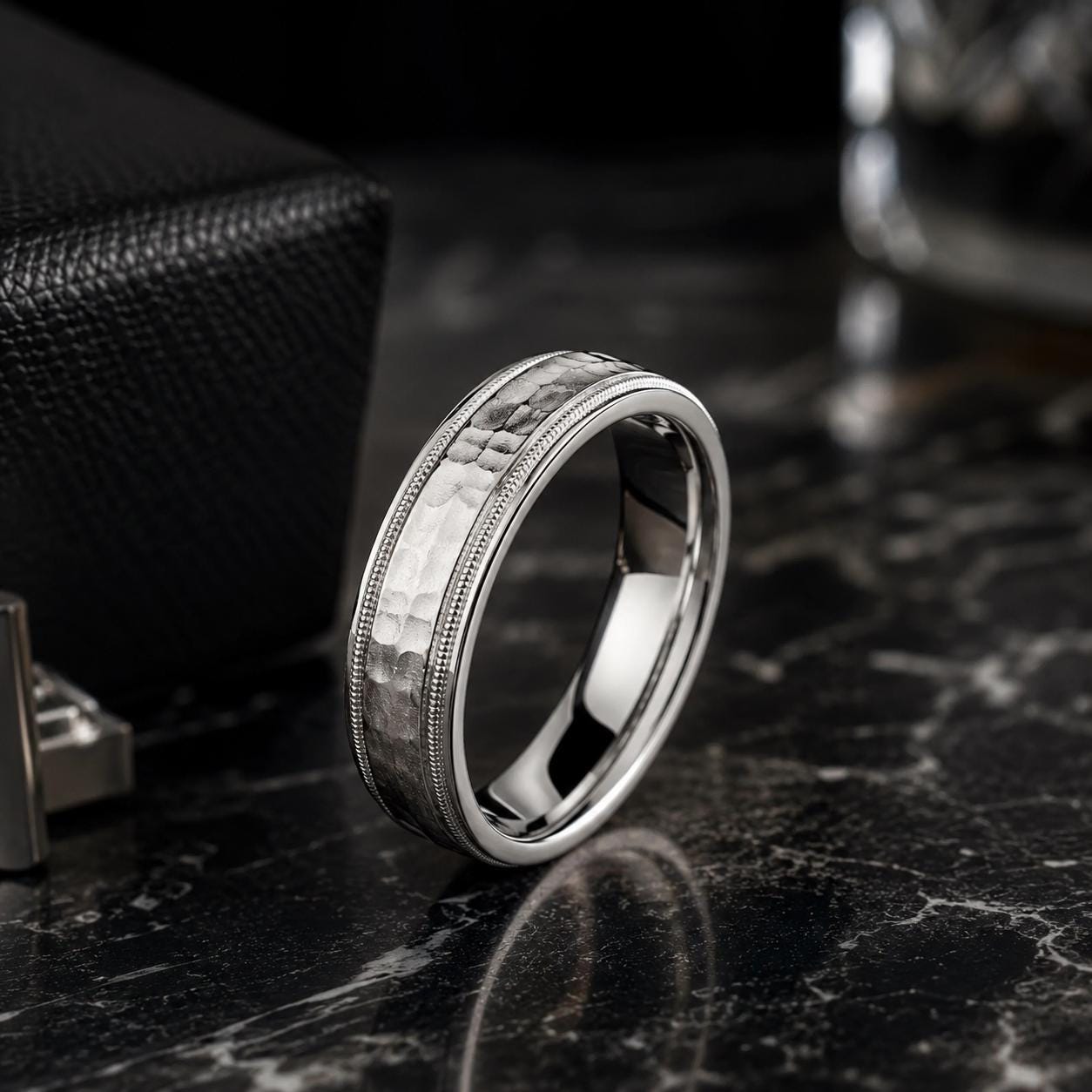 A silver ring with a textured band sits on a black marble surface, with a black leather box and a glass of water in the background.
