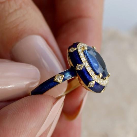 A close-up image of a person's hand holding a blue and gold ring with a large blue gemstone.