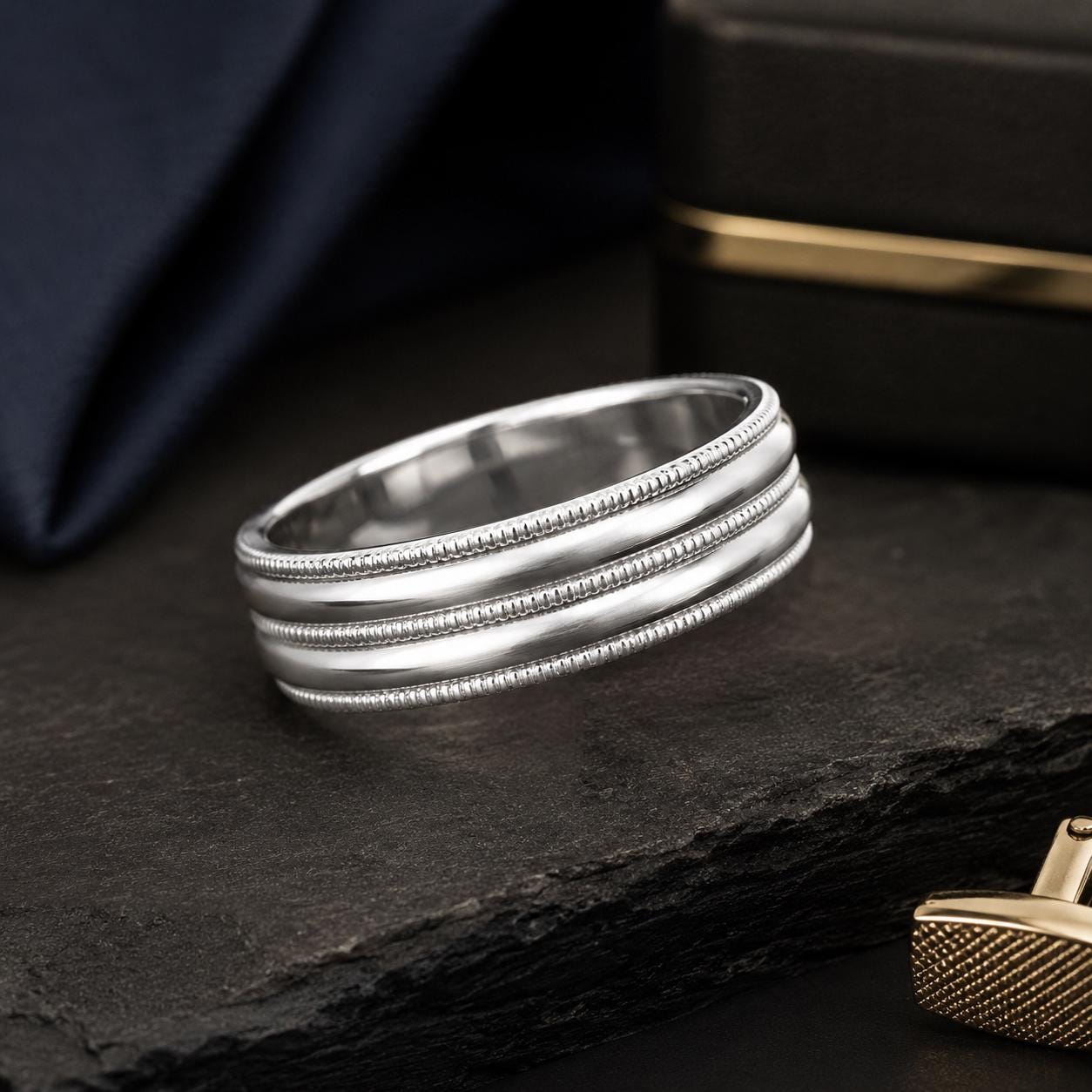 A silver ring with three rows of small, decorative beads is displayed on a dark surface, with a gold object partially visible in the background.