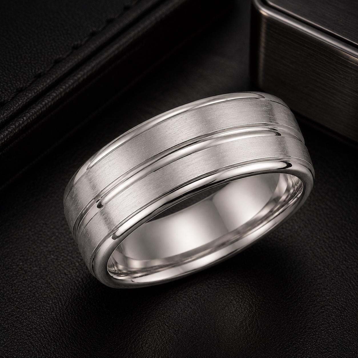 A silver ring with a textured band is displayed on a black surface, with a black leather-like object in the background.