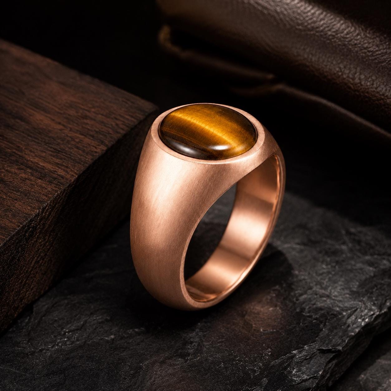 A gold-colored ring with a large amber-colored gemstone, possibly a tiger eye, is displayed on a dark surface.