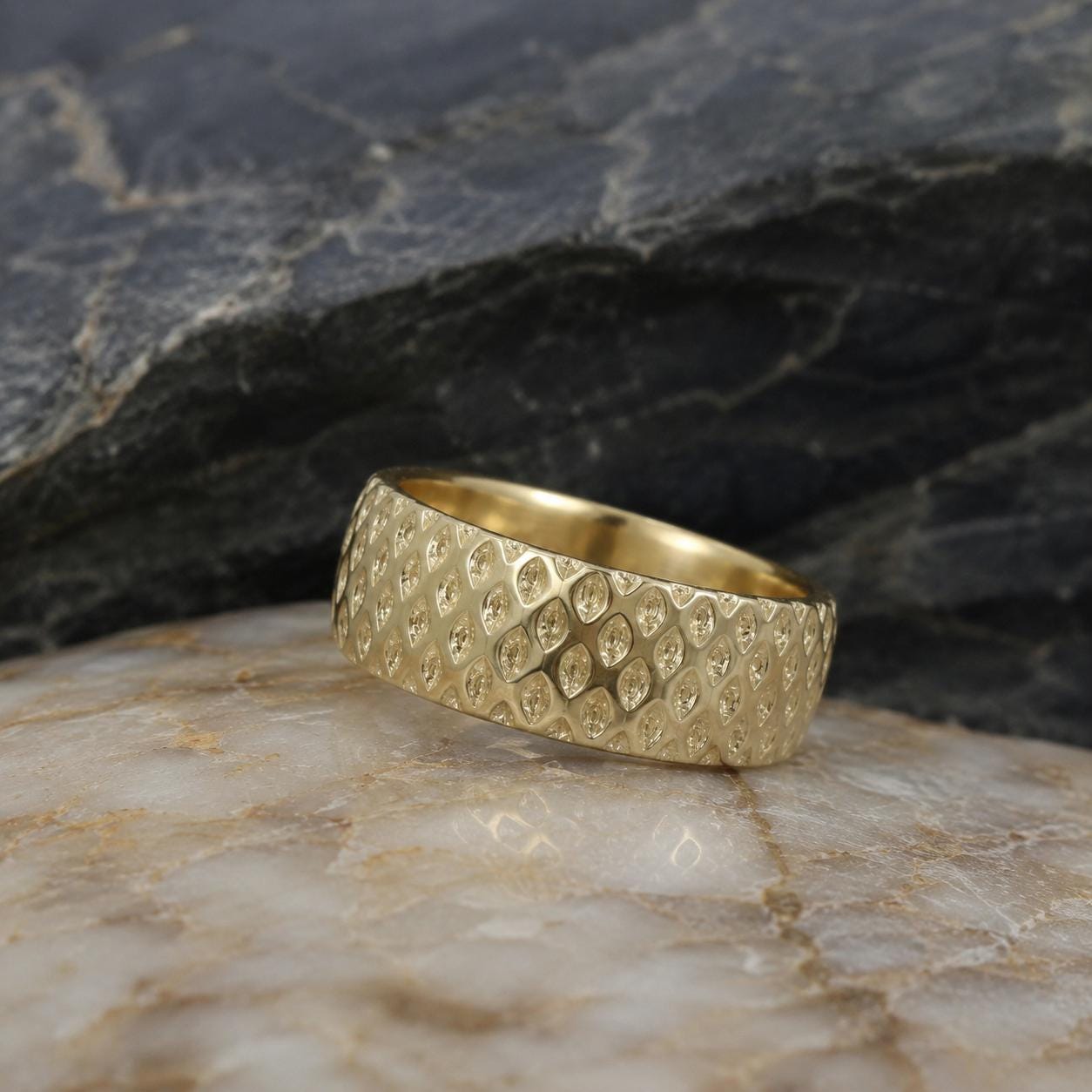 A gold ring with a textured surface sits on a marble surface, with a dark background.