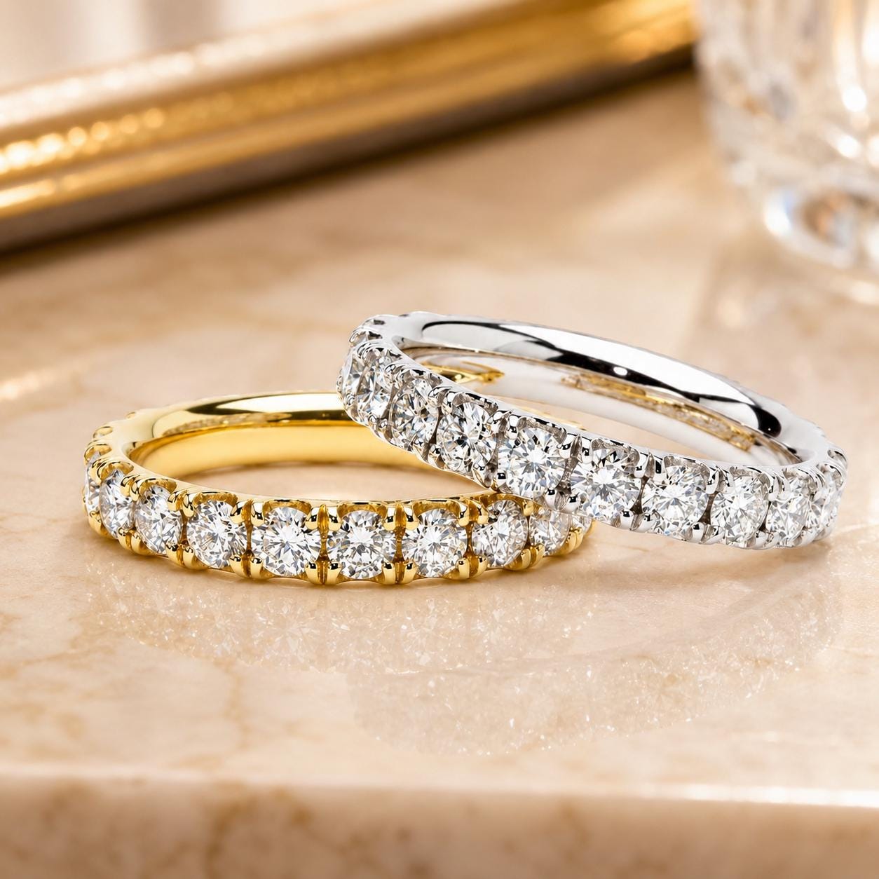Two diamond-encrusted rings are displayed on a marble surface, one in gold and the other in silver.
