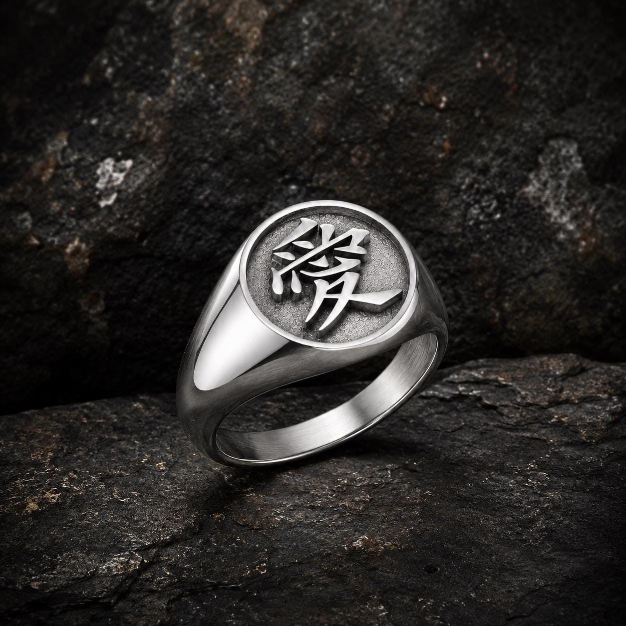 A silver ring with a circular design featuring an Asian character, likely a symbol or deity, is displayed against a dark, rocky background.