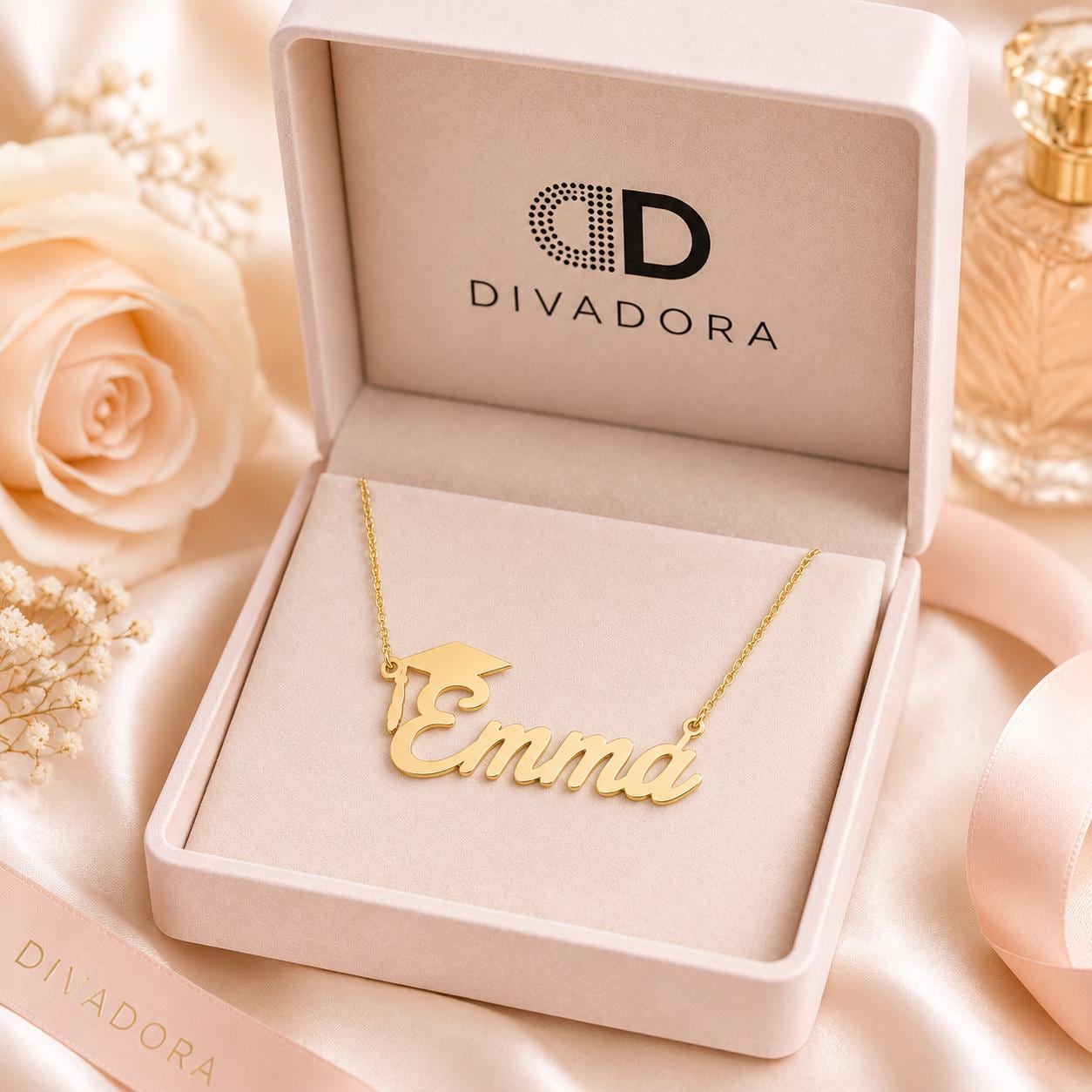 A gold necklace with the name "Emma" engraved on it, displayed in a white jewelry box with the Divadora logo. The necklace is placed on a pink satin background, with a rose and a perfume bottle visible in the background.