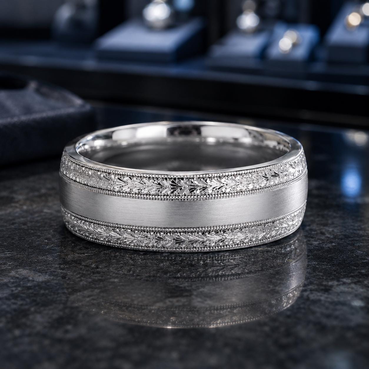 A silver ring with a decorative band sits on a dark surface, with a blurred background that appears to be a jewelry display.